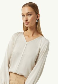 Beige langwerpige blouse met lange mouwen en een V-hals, gemaakt van een gladde stof. Heeft een sluiting aan de voorkant en licht geplooide mouwen.