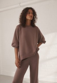 Felpa marrone oversize con maniche raglan, texture morbida e ampio girocollo, abbinata a pantaloni largi coordinati. Finitura del tessuto liscia.
