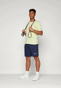 Gai zaļš īsām piedurknēm T-krekls, tumši zili šorti ar logo; modelis tur melnu lecamaukla, valkājot baltus sporta apavus un potīšu zeķes.