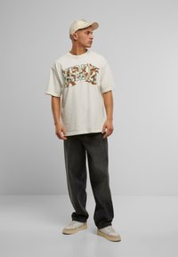 Off-white oversized T-shirt med farverig grafisk design. Mørke, løse jeans og beige sneakers. Lys cap fuldender det afslappede look.