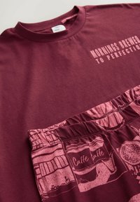 Burgundy puuvillapaita, jossa on valkoinen "Aamukahvi täydellisesti valmistettuna" -teksti; yhdistettynä vaaleanpunaisiin kuviollisiin shortseihin, joissa on kahviteemoisia kuvastoja.