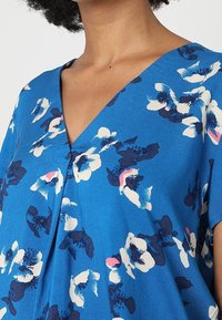 Blue short-sleeve top with a V-neck design, featuring a floral pattern in white, navy, and pink. Made from lightweight fabric, relaxed fit.