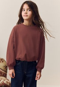 Viininpunainen collegepaita pyöreällä pääntiellä ja hieman liehuvilla hihoilla, yhdistetty tummansinisiin farkkuihin. Pehmeä kangas, rento istuvuus.