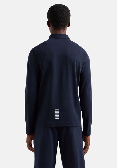 Homme vu de dos portant un haut bleu marine à manches longues et un pantalon avec un petit détail rayé blanc vertical près du bas du dos.