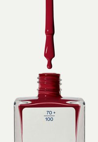 Smalto per unghie rosso in una bottiglia di vetro trasparente con una forma arrotondata. La bottiglia ha un'etichetta bianca con testo blu che riporta "70 + 100". Una goccia di smalto pende dal pennello.