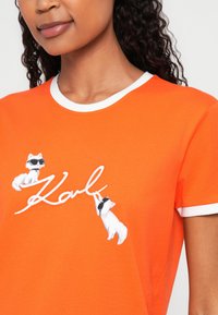 Oranžinė medvilninė marškinė su balta išribiota apykakle, vaizduojančios du animacinius katinus su akinių nuo saulės ir siuvinėtu "Karl" logotipu rašytiniu šriftu.