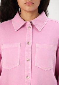 Pink denim shirt with a classic collar, front button closure, two chest pockets, and subtle textured fabric.