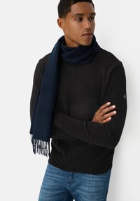 Navyblauer Wollschal, der um den Hals drapiert ist und fransige Kanten hat. Über einem strukturierten schwarzen Pullover getragen, mit rundem Halsausschnitt und gerippten Bündchen.