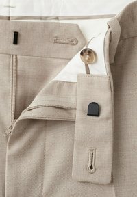 Vita dei pantaloni beige con bottone aperto, cerniera e dettaglio di chiusura a gancio e occhiello neri.