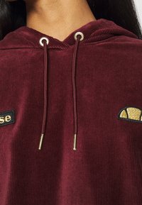 Bordeaux corduroy hoodie met zilveren oogjes, verstelbare koordjes en een zwarte patch met geborduurde tekst en een gouden grafisch accent.