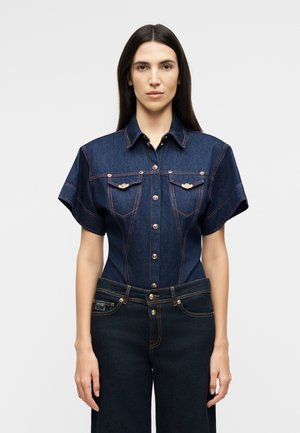 Γυναίκα με μακριά σκούρα μαλλιά, φοράει κοντομάνικο εφαρμοστό πουκάμισο από σκούρο τζιν και σκούρο τζιν παντελόνι, στέκεται μπροστά σε ένα απλό φόντο.