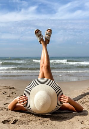 Szeroki kapelusz słomkowy w czarne paski, białe sandały z dwoma klamrami, ułożony na piaszczystej plaży z uniesionymi nogami i oceanem w tle.