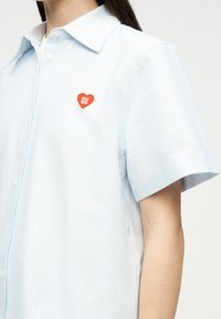 Άτομο που φοράει κοντομάνικο πουκάμισο με κουμπιά, ριγέ σε ανοιχτό μπλε και λευκό χρώμα, με ένα μικρό κόκκινο έμβλημα σε σχήμα καρδιάς που γράφει "HUGO" στο στήθος.