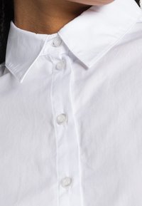 Camisa blanca de algodón con cierre de botón en la parte delantera, cuello clásico y textura suave. Presenta cinco botones redondos de plástico a lo largo de la tapeta.