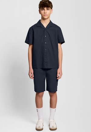 Teenager-Junge steht, trägt dunkles kurzärmeliges Hemd mit Knöpfen, passende Shorts, weiße Sportsocken und beige-weiße Turnschuhe, vor einfarbigem Hintergrund.