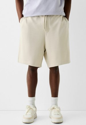 Shorts en coton beige avec une taille élastique et un cordon de serrage. Dotés de poches et d'une coupe décontractée, accompagnés de baskets blanches et de chaussettes.