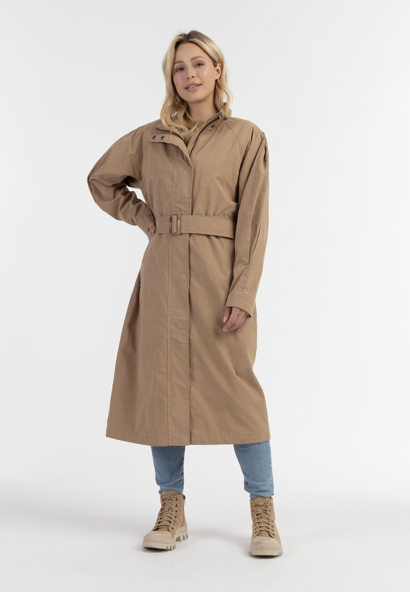 Béžový trench coat po kolena s velkými kapsami, páskem v pase a vysokým límcem; nošený přes světle modré džíny a hnědé kotníkové boty.