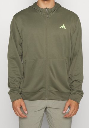 Person trägt einen olivgrünen Hoodie mit Reißverschluss und Kapuze sowie ein hellgrünes Logo auf der linken Brust, kombiniert mit hellgrauen Hosen.