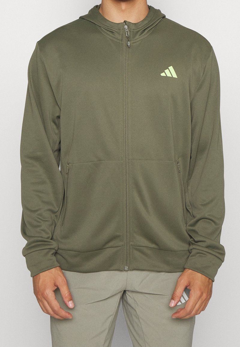 Personne portant un sweat à capuche zippé vert olive avec capuche et logo vert clair sur la poitrine gauche, associé à un pantalon gris clair.