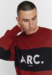 Rød og svart hettegenser med lange ermer, med en bred horisontal stripe og stor tekst "ARC." Enkel, rund halslinje og myk tekstur.
