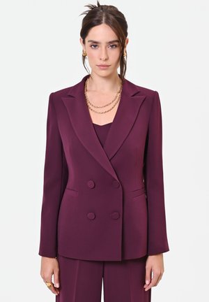 Donna che indossa un blazer doppiopetto color prugna e pantaloni abbinati, con una collana a catena dorata e orecchini, in piedi davanti a uno sfondo bianco.