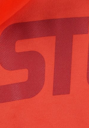 Czerwona tkanina o gładkiej fakturze z dużymi, ciemnoczerwonymi literami częściowo nakładającymi się na kolor tła, tworząc wyraźny wizualny kontrast.