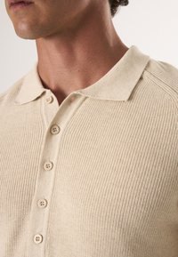 Beżowa dzianinowa koszulka polo z krótkim kołnierzykiem i pięcioma guzikami. Żebrowana struktura i dopasowany krój widoczne wokół szyi i ramion.