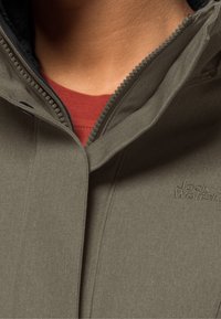 Olivgrön jacka med hög krage och blixtlås, med en strukturerad tyg och minimal branding på framsidan. En röd skjorta syns under.