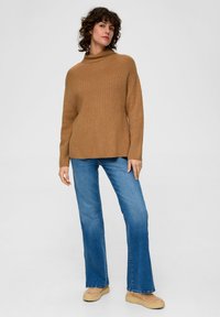 Brauner Strickpullover mit hohem Kragen, kombiniert mit hellblauen, ausgestellten Jeans und beigen Schuhen, vor einem neutralen Hintergrund.