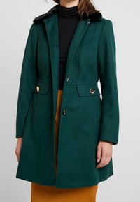 Cappotto verde in misto lana con colletto in pelliccia sintetica nera, hardware dorato, silhouette sartoriale e tasche a soffietto. Texture liscia.