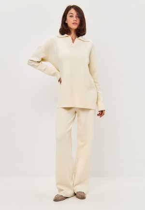 Jersey beige con cuello en V, combinado con pantalones de pierna ancha a juego. Textura suave y corte relajado. El calzado es de color marrón claro.