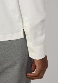 Camicia bianca a maniche lunghe con polsini abbottonati e dettagli di cucitura, abbinata a pantaloni grigi con texture, che mostrano un contrasto di tessuti.