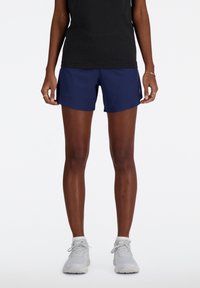 Pantaloni corti sportivi blu navy con una texture liscia, caratterizzati da un piccolo logo sul lato, indossati con una canotta nera e sneakers di colore chiaro.