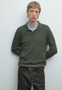 Ung mand med blondt hår iført mørkegrøn ribbet sweater og grå skjorte, stående med hænderne bag ryggen mod en ensfarvet hvid væg.