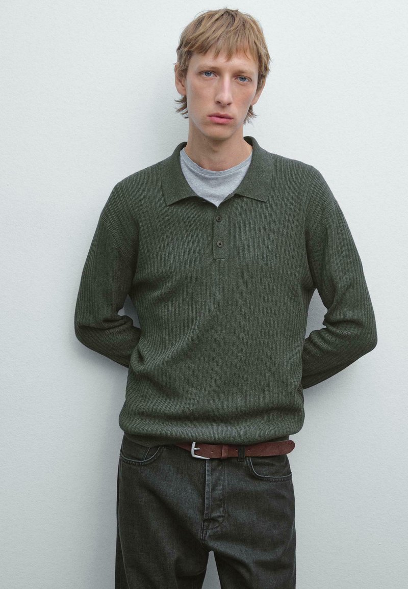 Ung mand med blondt hår iført mørkegrøn ribbet sweater og grå skjorte, stående med hænderne bag ryggen mod en ensfarvet hvid væg.