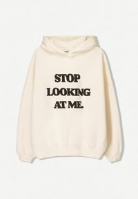 Béžová mikina s kapucí a černým tučným textem "PŘESTAŇ SE NA MĚ DÍVAT." na přední straně, vystavená na bílém pozadí.