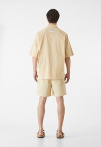 Tričko s krátkym rukávom svetložltej farby v kombinácii s ním vhodnými šortkami. Hladká tkanina, voľný strih a štítok na zadnej strane. Celkový vzhľad dopĺňajú sandále.