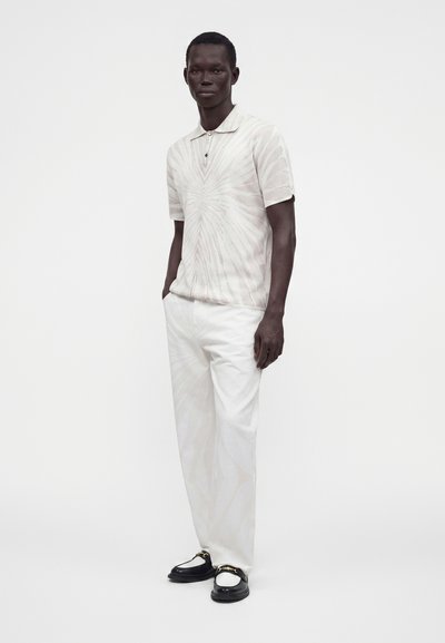 Homme debout portant un polo tie-dye clair, un pantalon blanc et des mocassins noir et blanc avec un détail chaîne dorée.