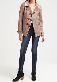 Brun ruskindsjakke med krave af kunstpels, togglesløsninger og brede slag, parret med mørke denimjeans og sorte ankelstøvler.