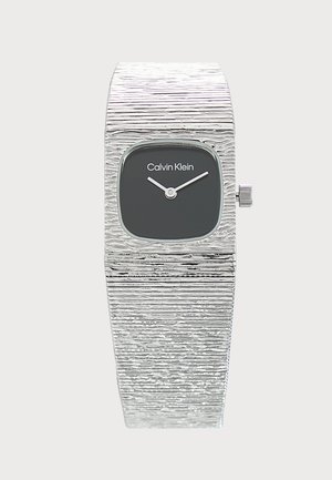 Srebrny teksturowany zegarek na rękę Calvin Klein z małą czarną kwadratową tarczą i dwoma prostymi białymi wskazówkami pokazującymi godzinę 10:10.