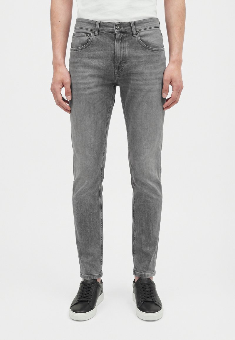 Homme portant un jean gris slim, des baskets noires et une chemise blanche, debout devant un fond blanc uni.