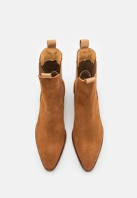 Stivali alla caviglia in suede marrone con punta affusolata, design senza lacci e piccole linguette sui lati. Fodera interna liscia e suola in pelle.