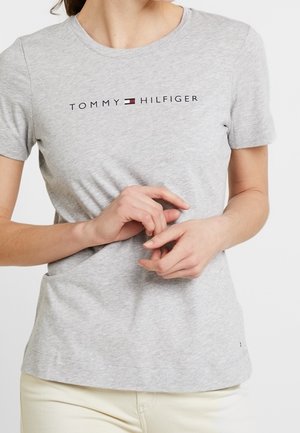 Personne portant un T-shirt gris clair à manches courtes avec le logo "Tommy Hilfiger" et un pantalon de couleur crème, mains délicatement jointes devant.