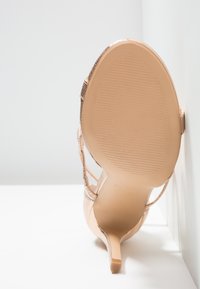 Chaussure beige à talon haut avec semelle texturée, fine bride et talon pointu, positionnée à la verticale contre un fond blanc et un mur.