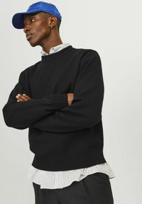 Schwarzer Strickpullover mit Rundhals-Ausschnitt, über einem weiß-blau gestreiften Hemd getragen. Eine blaue Mütze und schwarze Hosen runden das Outfit ab.
