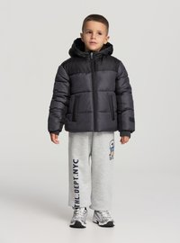 Kurtka puchowa w kolorach czarnym i szarym, zapinana na zamek, z kapturem i elastycznymi mankietami. Noszona ze szarymi dresami i trampkami.