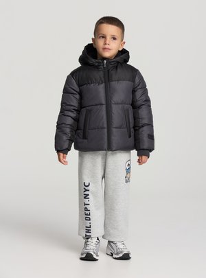 Giacca imbottita, design a blocchi di colore nero e grigio, chiusura con zip, cappuccio, polsini elastici. Indossata con pantaloni della tuta grigi e sneakers.