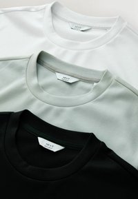 Tres camisetas de cuello redondo en blanco, gris claro y negro. Material de algodón suave con cuellos y etiquetas en contraste que indican la marca y el ajuste.