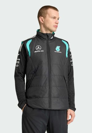 Man wearing black padded racing vest with turquoise accents and Mercedes AMG Petronas logos, looking right with hands in pockets.