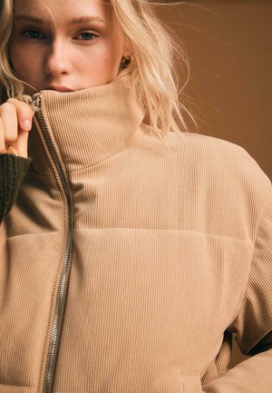 Beige fløjljakke med høj krave, synlig lynlås og ribbet tekstur. Syning detaljer tilfører dybde til designet.